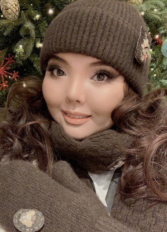 Een vrouw met een muts, sjaal en wanten, met een kerstboom op de achtergrond.