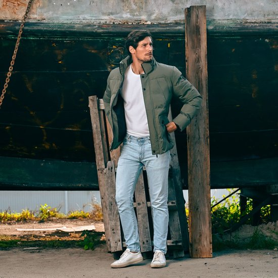 Man met olijfgroene winterjas, witte t-shirt en lichtbruine spijkerbroek, staand bij een boot.