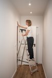 Een vrouw staat op een aluminium Baulich trap in een gang en monteert een gordijnroede aan de muur.