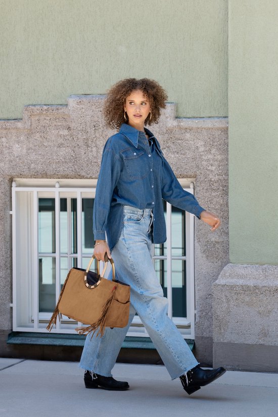 Model loopt met een bruine suède tas met franjes en gouden handvatten. Ze draagt een spijkerblouse en jeans.