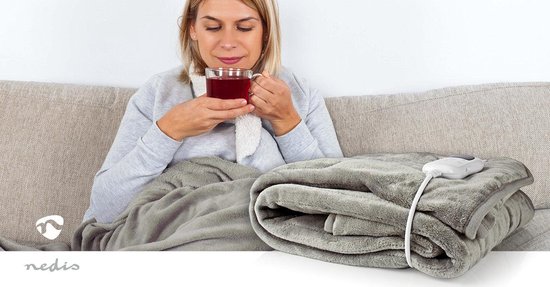 Grijze fleece elektrische deken, gevouwen, met bedieningskabel en afstandsbediening. Vrouw drinkt thee.