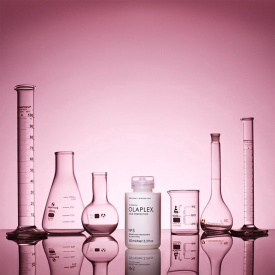 Een fles Olaplex Hair Perfector No.3 staat tussen laboratoriumglaswerk tegen een roze achtergrond.