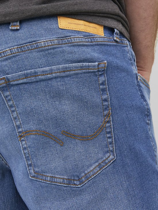 Blauwe jeans met een gele leren label met de tekst 'CRAFTSMANSHIP AND DEDICATION'. Oranje stiksels op de achterzakken.