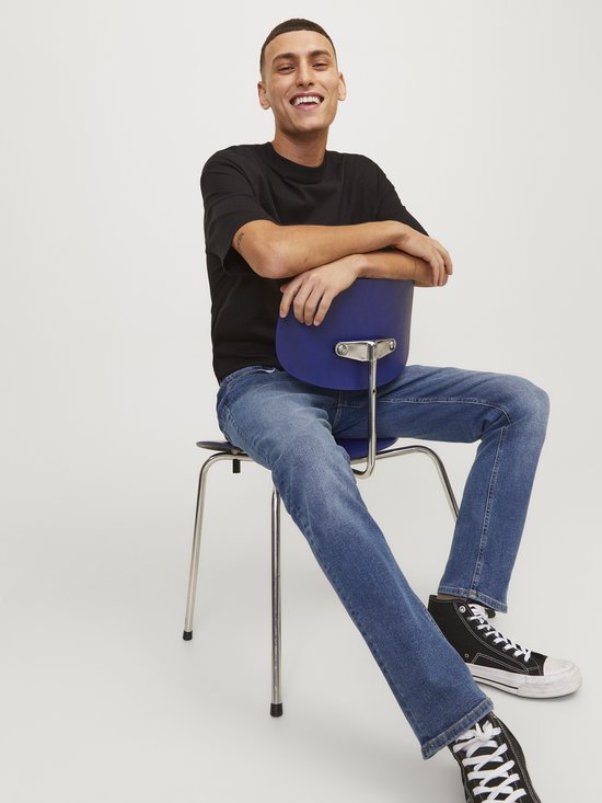 Model zit op een stoel, gekleed in een zwart T-shirt en loszittende spijkerbroek.