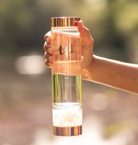 Hand houdt een glazen fles met koperkleurige dop en rozenkwarts kristallen vast.