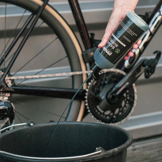Hand die DYNAMIC BADASS BUBBLES fles leegt in zwarte emmer, naast fiets.