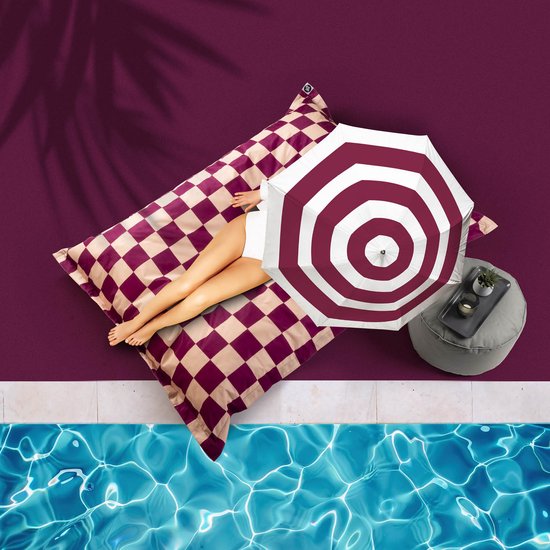 Vrouw met benen op paars-crèmekleurig dambord ligkussen bij zwembad, onder parasol.