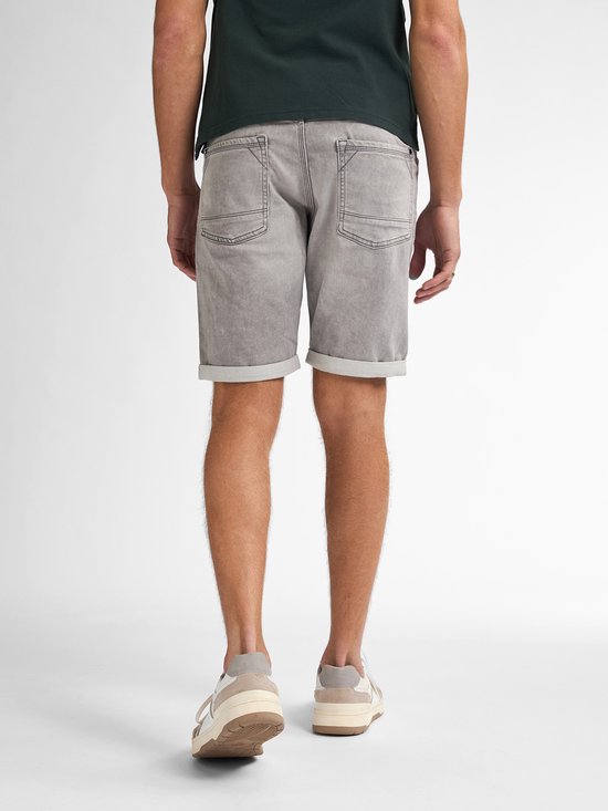 Man van achteren gezien, draagt een donkergroene polo en een grijze denim short met opgerolde zomen. Hij draagt witte sneakers.