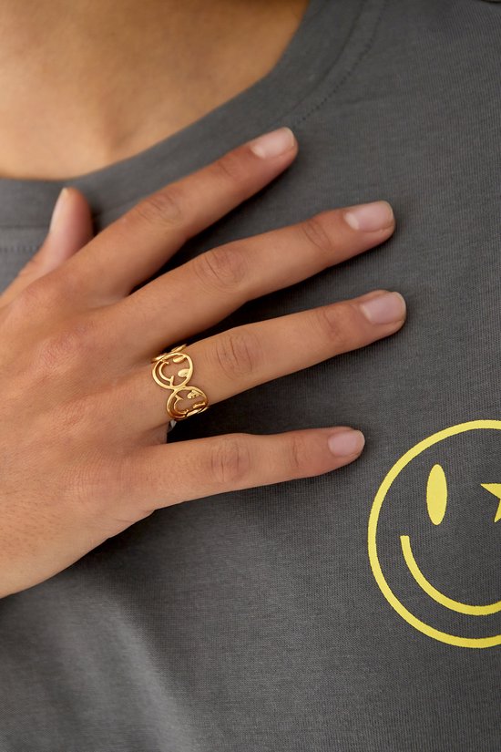 Gouden ring met meerdere smiley gezichten, gedragen op een hand op een grijs T-shirt.