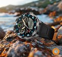 Zilveren smartwatch met zwart, geperforeerd bandje, op rotsen bij de zee, display toont tijd en stappenteller.