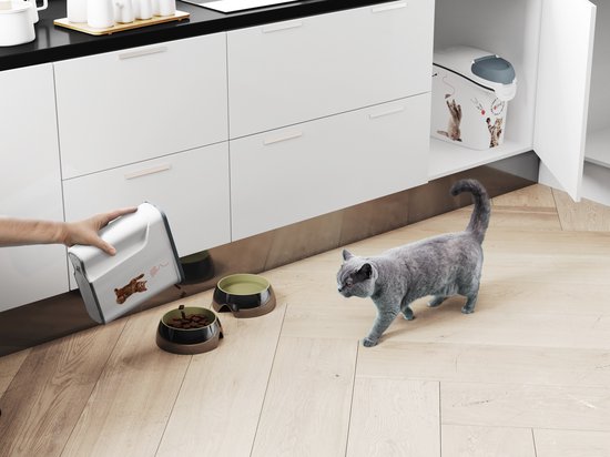 Witte voedselcontainer met kattenafbeeldingen, onder een keukenkastje, met een kat op de vloer.