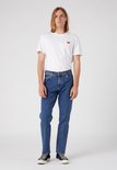 Man met lang blond haar, staand, in blauwe spijkerbroek en wit t-shirt met logo.