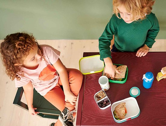 Deux enfants à table avec des boîtes à lunch et des gourdes. Un garçon en pull vert boit.