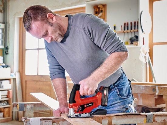 Man met baard in grijze trui en spijkerbroek zaagt met een rode Einhell decoupeerzaag een houten plank.