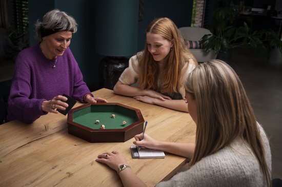 Drie vrouwen spelen een dobbelspel rond een achthoekige bak met groen vilt. Vijf dobbelstenen liggen in de bak.