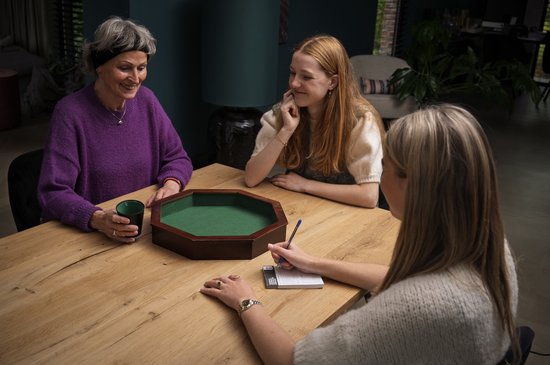 Drie vrouwen spelen een dobbelspel rond een achthoekige bak met groen vilt. Twee dobbelstenen liggen in de bak.