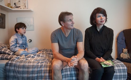 Man in grijs shirt en vrouw in zwart shirt zitten op een bed naast een jongen in een blauw gestreept pyjama.