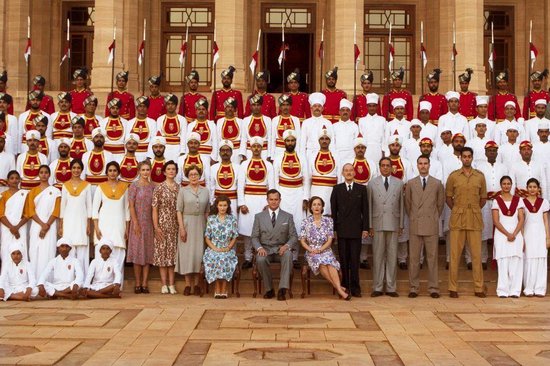 Groep mensen in traditionele kledij en westerse kleding voor een gebouw met rode vlaggen. De mannen in het midden dragen een ceremonieel uniform.