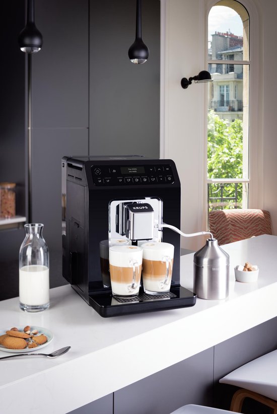 Zwarte Krups espressomachine op een keukenblad met twee glazen cappuccino en melkreservoir.