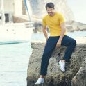 Man in donkerblauwe joggingbroek en geel T-shirt zit op rotsen bij het water.