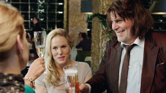 Een man met een bruin pak en een vrouw met blond haar kijken elkaar aan en houden glazen omhoog in een restaurant.