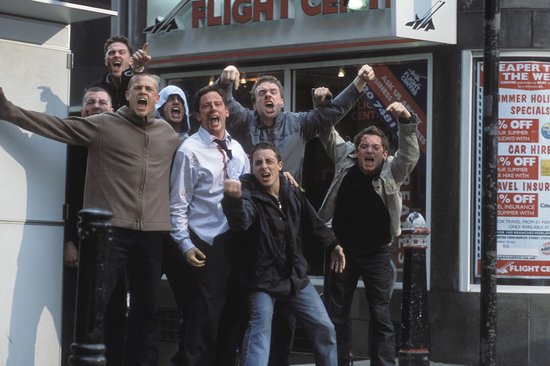 Een groep mannen schreeuwt en gebaart op straat voor een winkel met 'Flight Centre' en reclames voor vakanties.