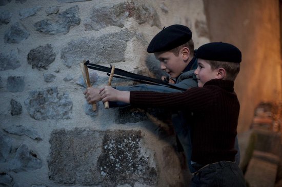 Twee jongens met baret richten met een katapult op een stenen muur.