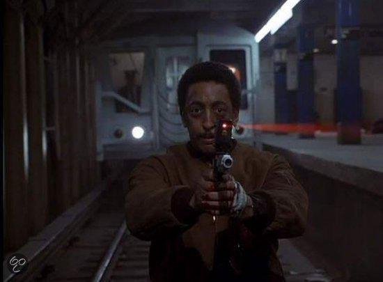 Man met snor en bruine jas richt een pistool met laser op de camera, in een metrostation.