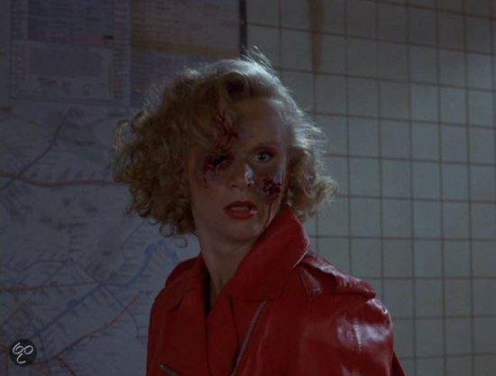 Vrouw met warrig blond haar, rode jas, en verwond gezicht met bloed.