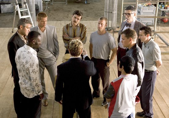 Groep van negen mannen die in een cirkel staan op een houten vloer, waarschijnlijk tijdens een filmopname.