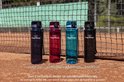Vier transparante drinkflessen in donkerblauw, roodpaars, blauw en zwart, met productcodes.