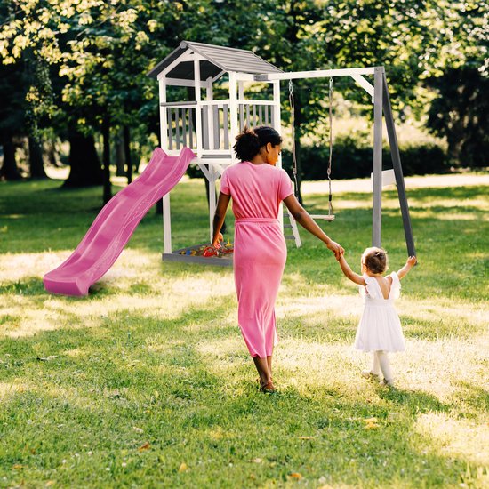 Moeder in roze jurk leidt haar dochter aan de hand naar een speeltoren met paarse glijbaan en schommel in een tuin.