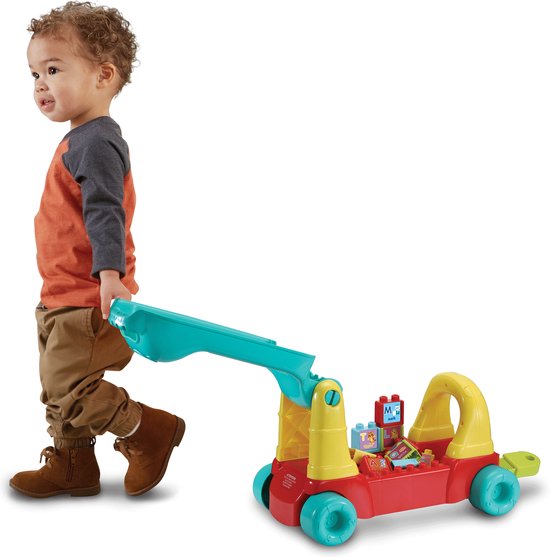 Een jongetje duwt een rode en gele loopwagen met een laadbak en blokken.