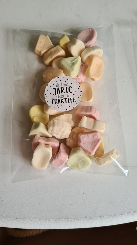 Een doorzichtig zakje gevuld met pastelkleurige snoepjes in de vorm van huisjes en andere figuren. Een ronde sticker met 'ik ben JARIG dus ik TRAKTEER' is zichtbaar.