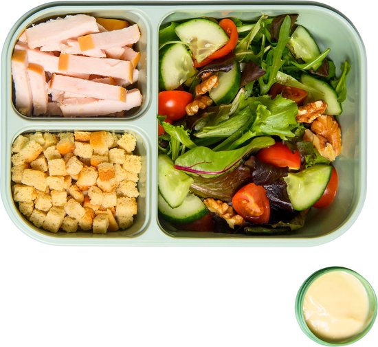 Groene lunchbox met drie vakken, gevuld met plakjes kalkoenfilet, croutons en een salade van sla, komkommer, cherrytomaatjes, rode ui en walnoten, met een bakje dressing.
