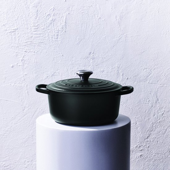 Zwarte Le Creuset braadpan met deksel en handvatten, tentoongesteld op een witte sokkel.