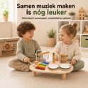 Twee kinderen spelen met een houten muziektafel met trommels, xylofoon, cimbalen en ratels. De tafel is omringd door cadeautjes en planten.