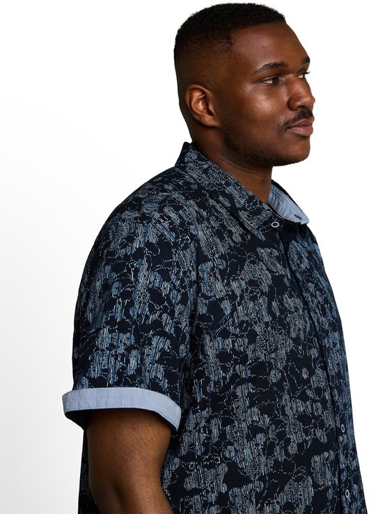 Model in een donkerblauw, kortemouwen overhemd met een abstract, lichtblauw en grijs patroon. De kraag en omgeslagen mouwen hebben een lichte denimkleur.