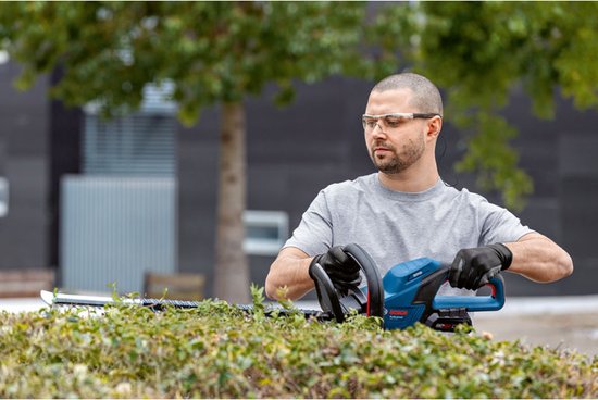 Man met blauwe Bosch Professional heggenschaar snoeit een groene, dichte struik.