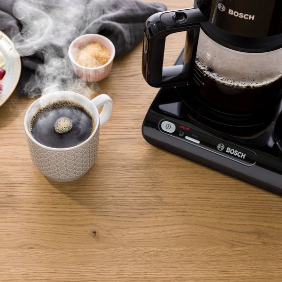 Zwarte Bosch koffiemachine op houten tafel, naast een mok dampende koffie en suiker.