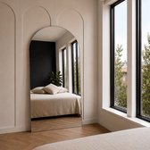 Grote boogspiegel in een lichte slaapkamer, weerspiegelt een bed met beige sprei en hoofdkussens, en twee grote ramen.