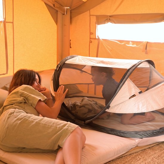 Een vrouw kijkt naar een kind in een wit en zwart Deryan campingbedje in een tent.