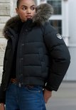 Detailopname van een zwarte korte gewatteerde jas met bontkraag en Superdry logo op de mouw, gedragen met spijkerbroek.