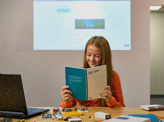 Een meisje met een oranje trui zit aan een tafel en leest een Arduino-projectenboek, met een laptop en elektronica op tafel.