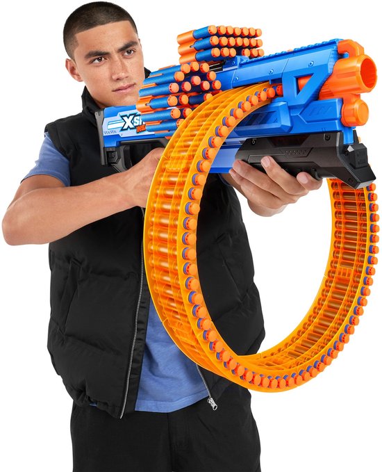 Een man met een zwarte bodywarmer en blauw shirt houdt een grote blauwe en oranje speelgoedblaster met een rond magazijn vast.