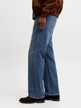 Man met de hand in de zak van een wijde blauwe jeans, zittend.