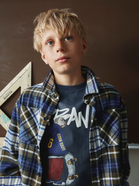 Jongen draagt een olijfgroen, blauw en wit geruit overshirt over een donkerblauw T-shirt met grafische print.