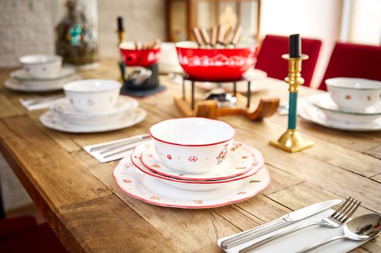 Gedekte tafel met kerstservies, waaronder borden en kommen met kerstafbeeldingen, rode fonduepan en gouden kandelaar.