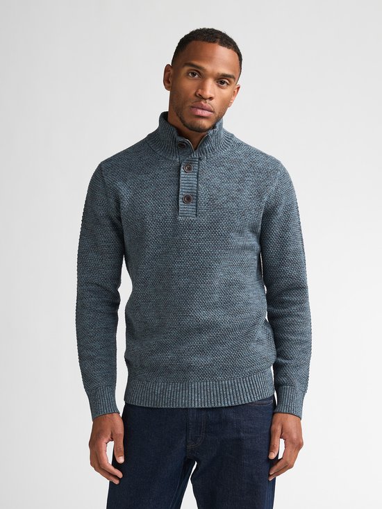 Man draagt blauwe gemêleerde sweater met hoge kraag en knoopjes, met donkerblauwe jeans.