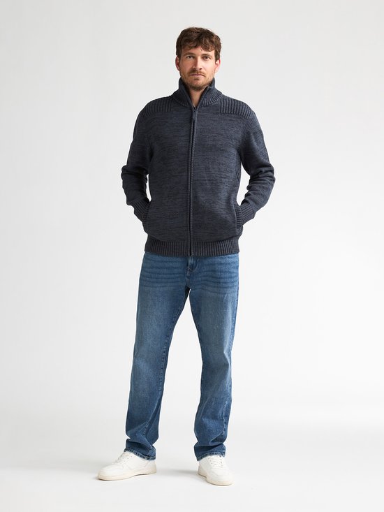 Man draagt donkerblauw gemêleerd gebreid vest met ritssluiting, hoge kraag, steekzakken en witte sneakers.